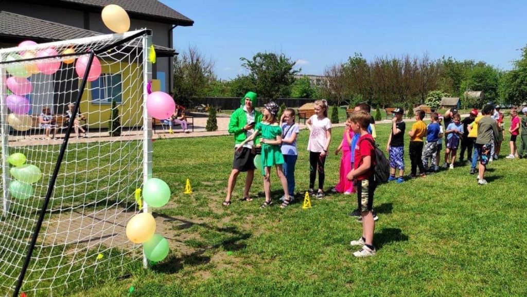 Children playing a game outside under blue skies in front of a football goal decorated with balloons