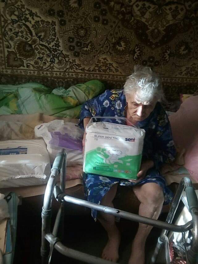 Old man sitting on a bed holding a pack of incontinence pads, with a walking frame in front of him