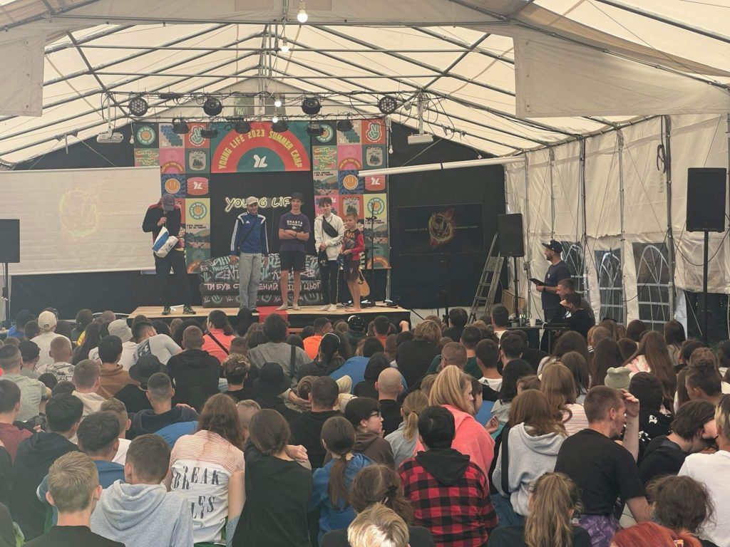 A leader and children standing on a stage in a marquee in front of other children seated on the floor of a marquee at the Young Life 2023 Summer Camp