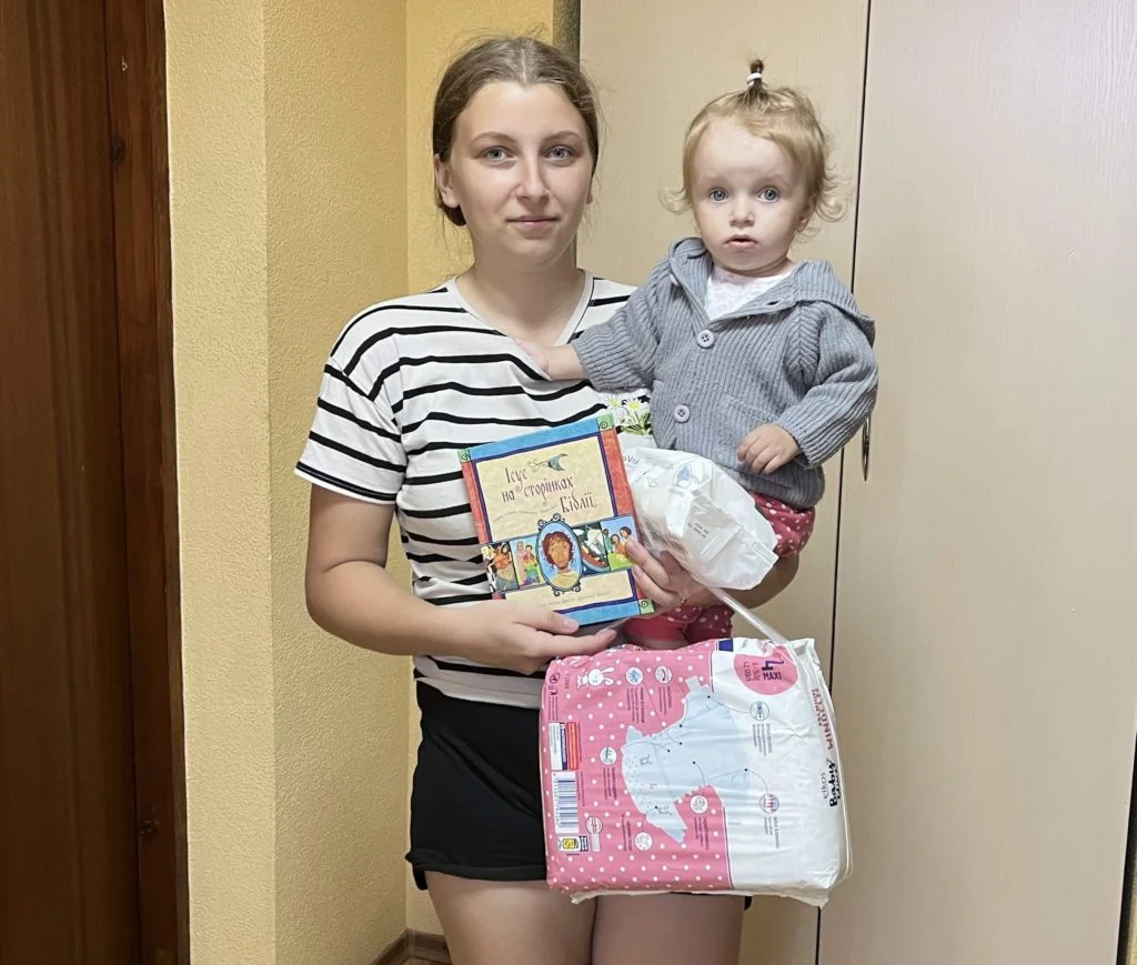 Mum holding her toddler and packs of nappies, baby wipes and a copy of the Jesus Storybook Bible
