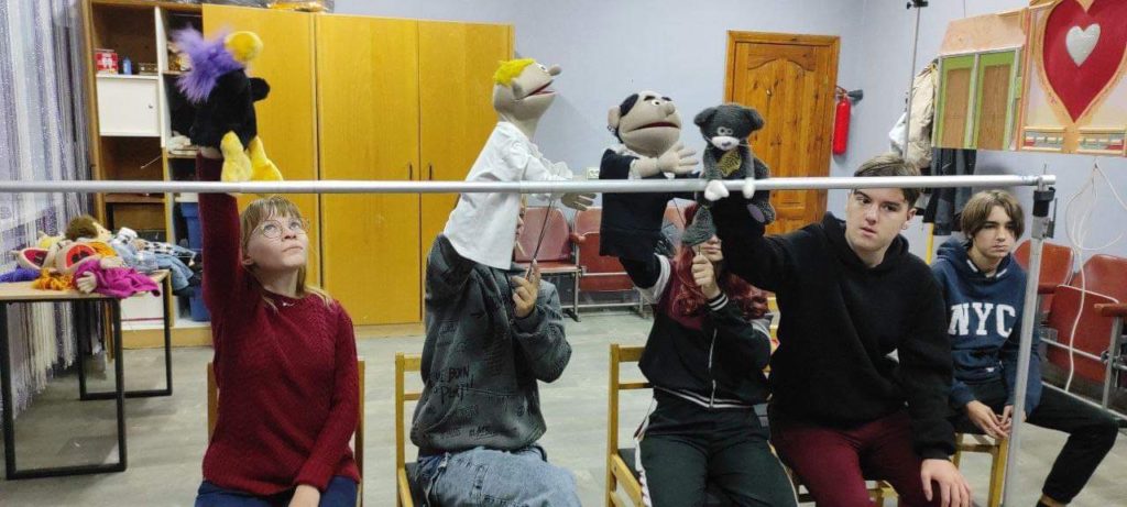 The puppet theatre team sitting behind the empty frame of their stage rehearsing their show