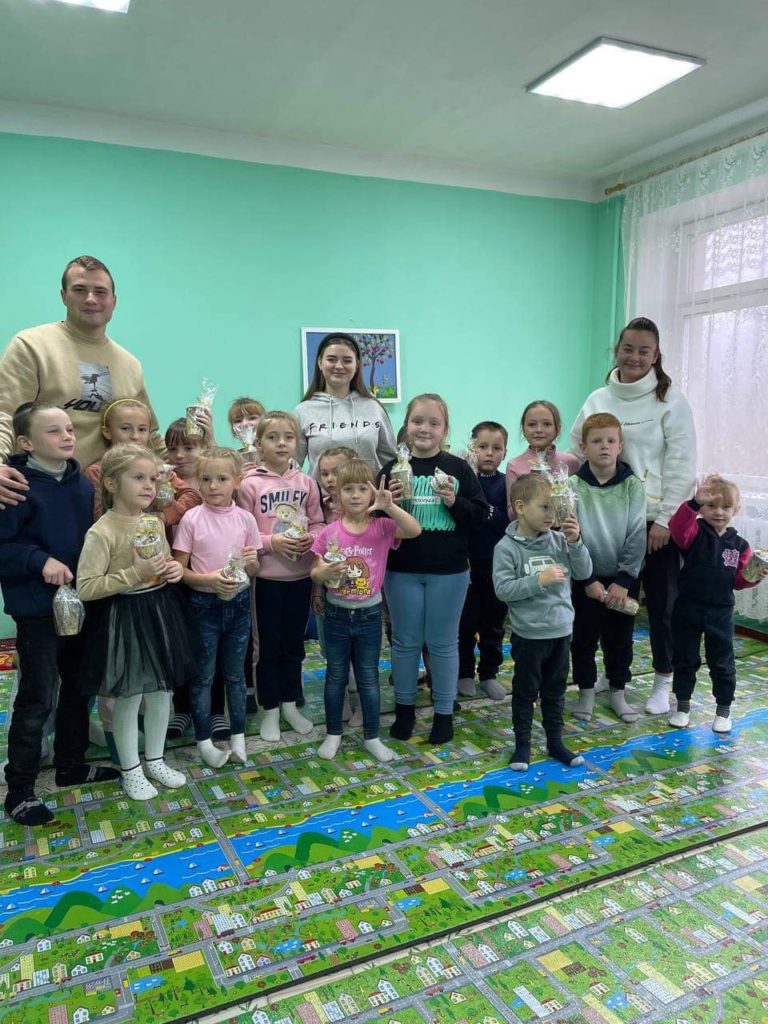 Valentine, Lyuba and Svitalana standing with a group of children at the local hospital in a room with a play mat on the floor featuring a cartoon village