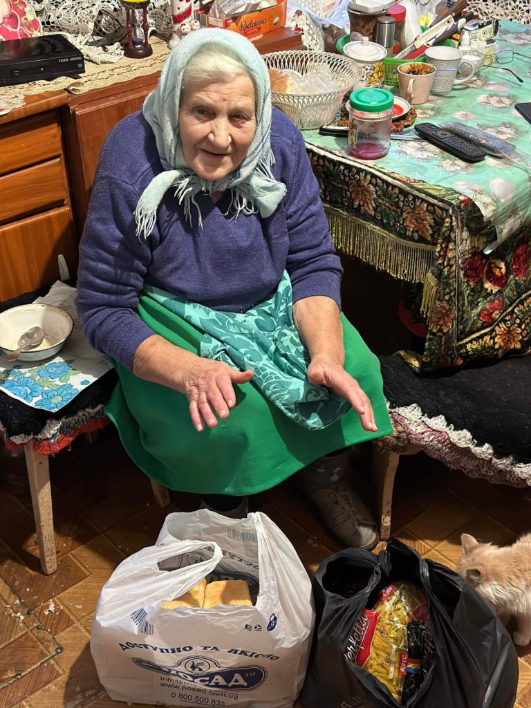 An elderly ladt sat in a chair in her home, with two bags of food aid on the floor in front of her