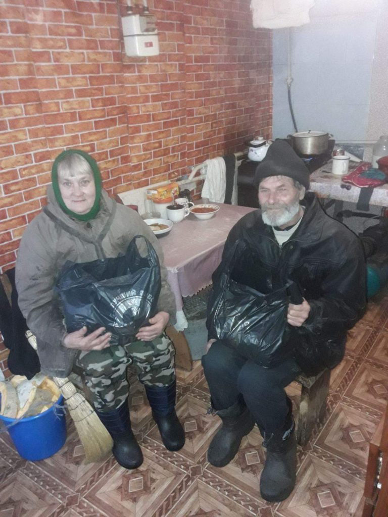 An elderly couple sat in a room each holding a bag of food aid