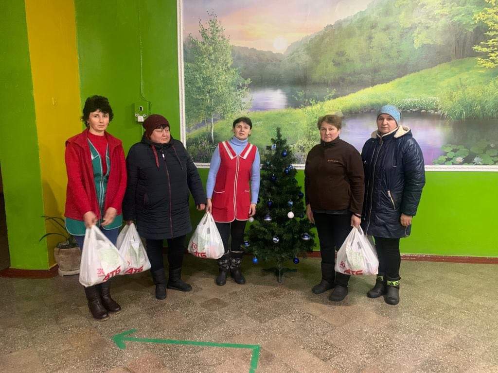 Five adults standing in a hall next to a small Christmas tree holding bags of aid