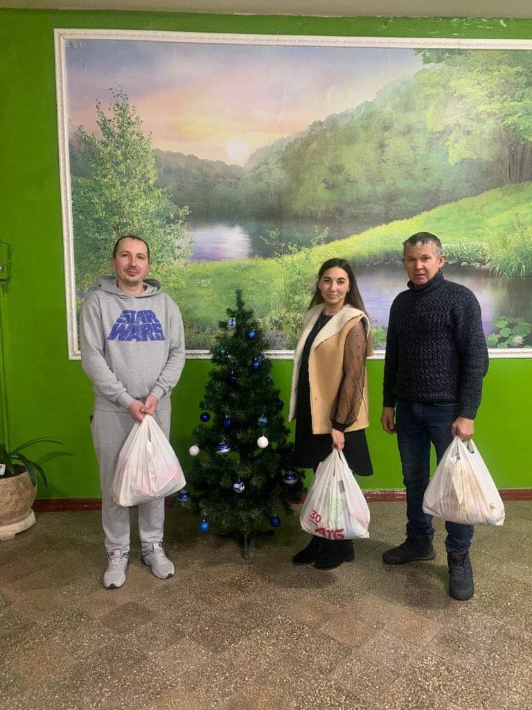 Three adults standing in a hall next to a small Christmas tree holding bags of aid