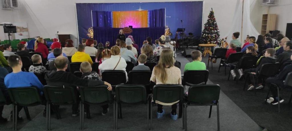 People sitting in the hall at the Centre during the Christmas Camp watching a child speak at the front.