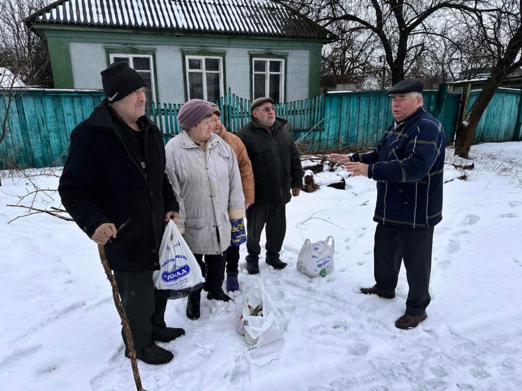 A group of elderly men and women standing in the snow with bags of food aid