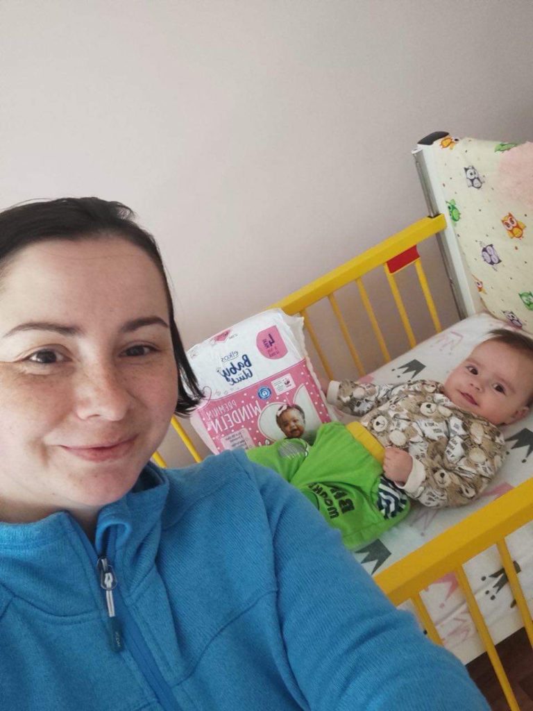 Lady taking a selfie with her baby lying in a cot behind her along with a pack of donated nappies