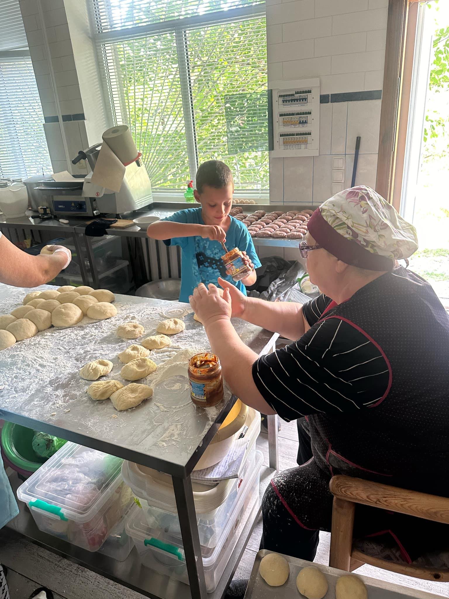 a lady sitting at a table in the bakery kneading dough, with a young boy standing at the table helping