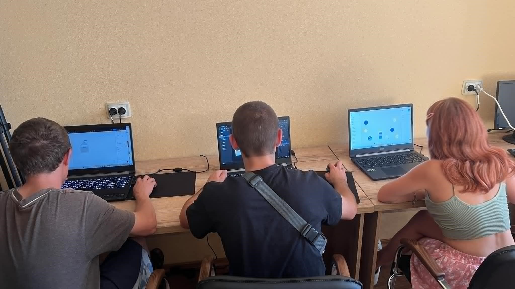 Boys and girls with their backs to us in a computer workshop sitting at a long bench looking at laptop screens