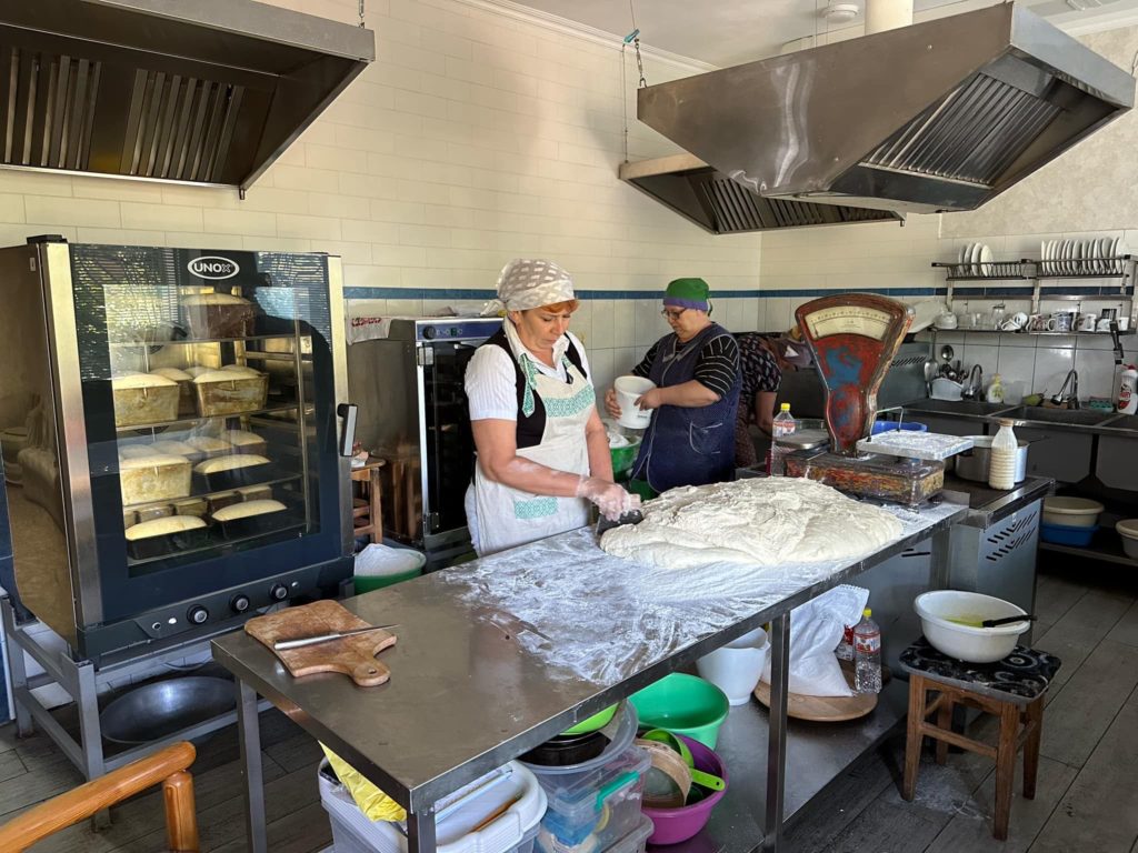 A large slab of dough spread out on a metal bench in the bakery with a lady cutting pieces off it ready for baking