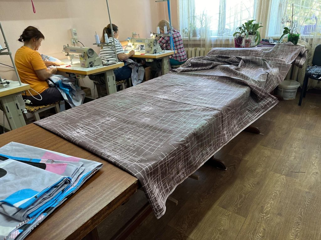 A large piece of material laid out on a table ready to be made into bedding, with three ladies sat at sewing machines in the background