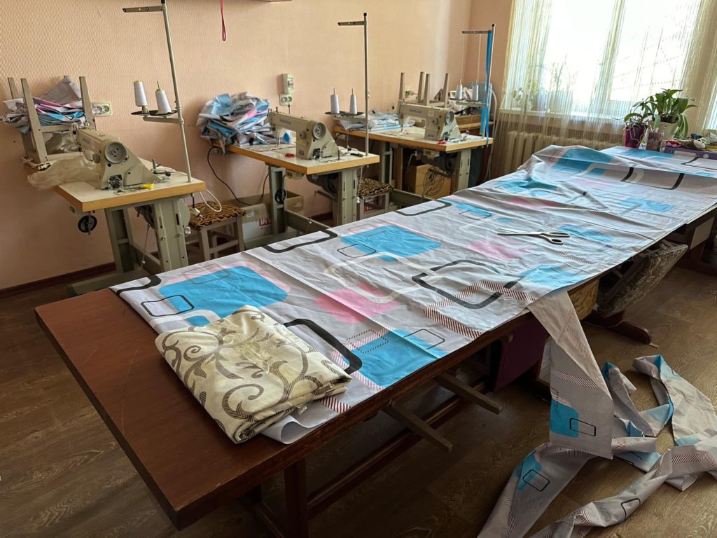 A large piece of material laid out on a table ready to be made into bedding, with three empty sewing machine stations in the background