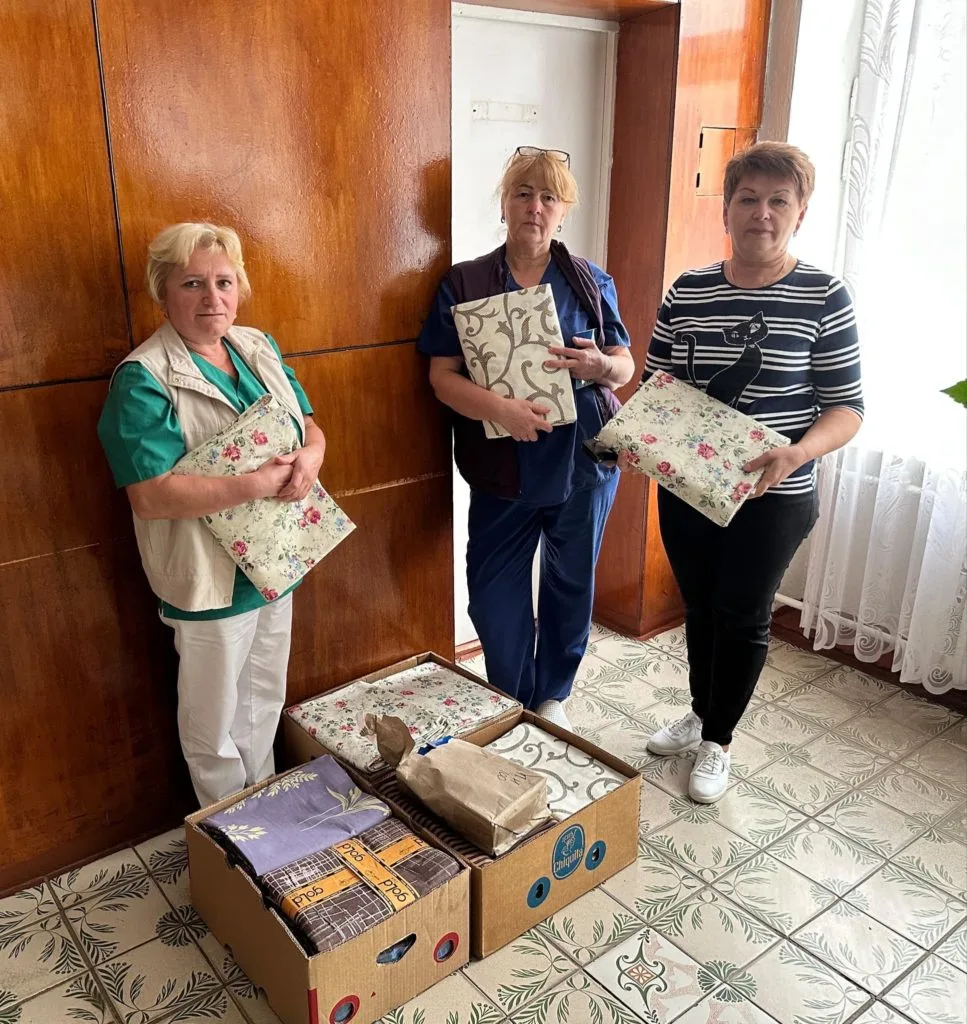 Three ladies standing holding bed linen that has been donated, with three boxes of the linen on the floor in front of them