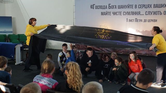 Children sat in chairs watching another group of children sat on the floor who are being covered in a large piece of black material