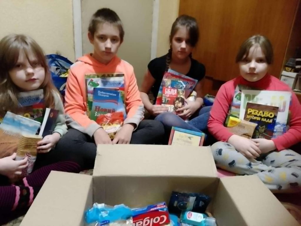 Children from a large family sitting on the floor around a box of donated toiletries, books, and food, holding some of those books