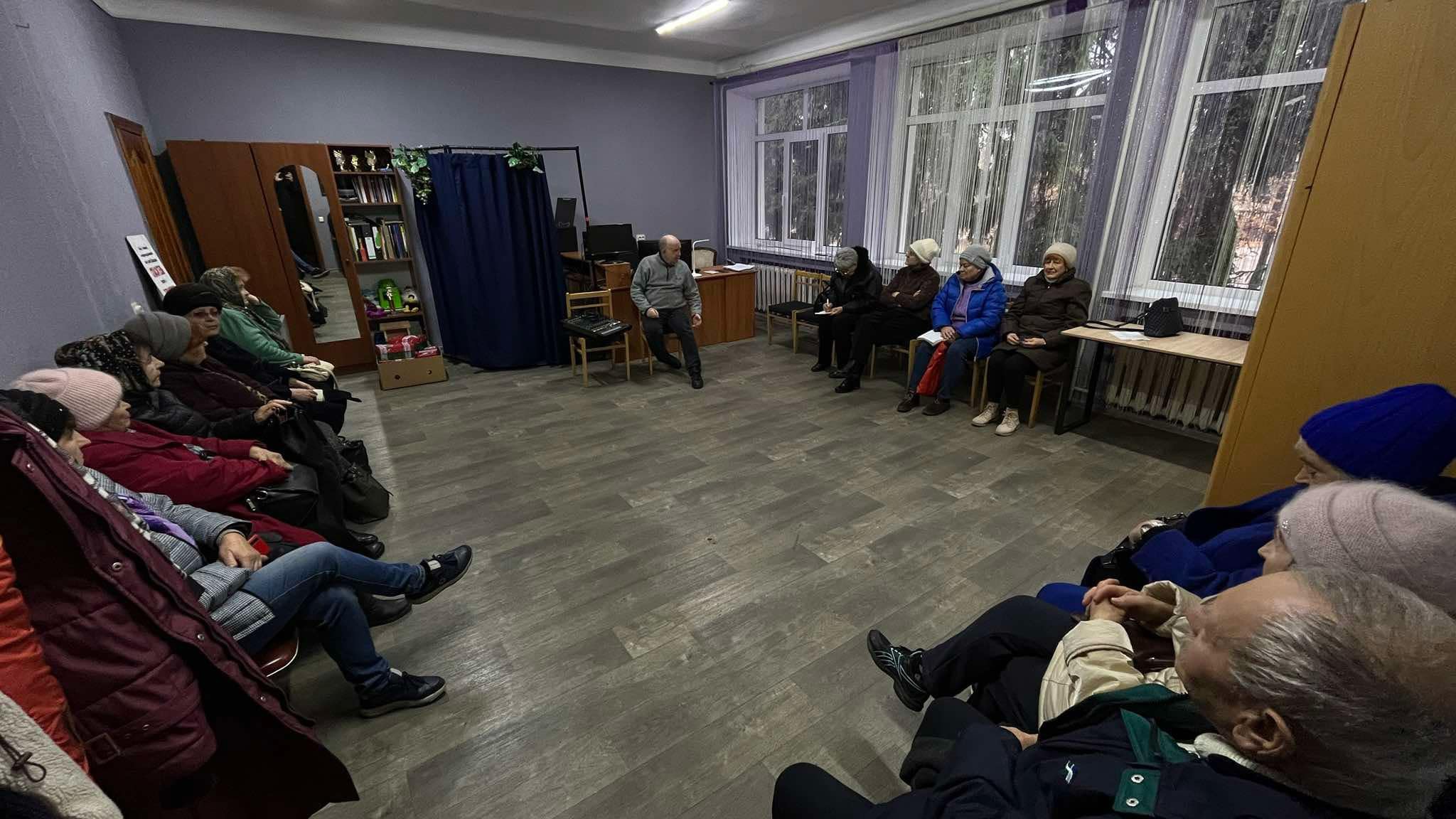 Musical Sergey sitting in a large room with a group of older adults sat around the edge, teaching them how to use smart phones and computers to communicate