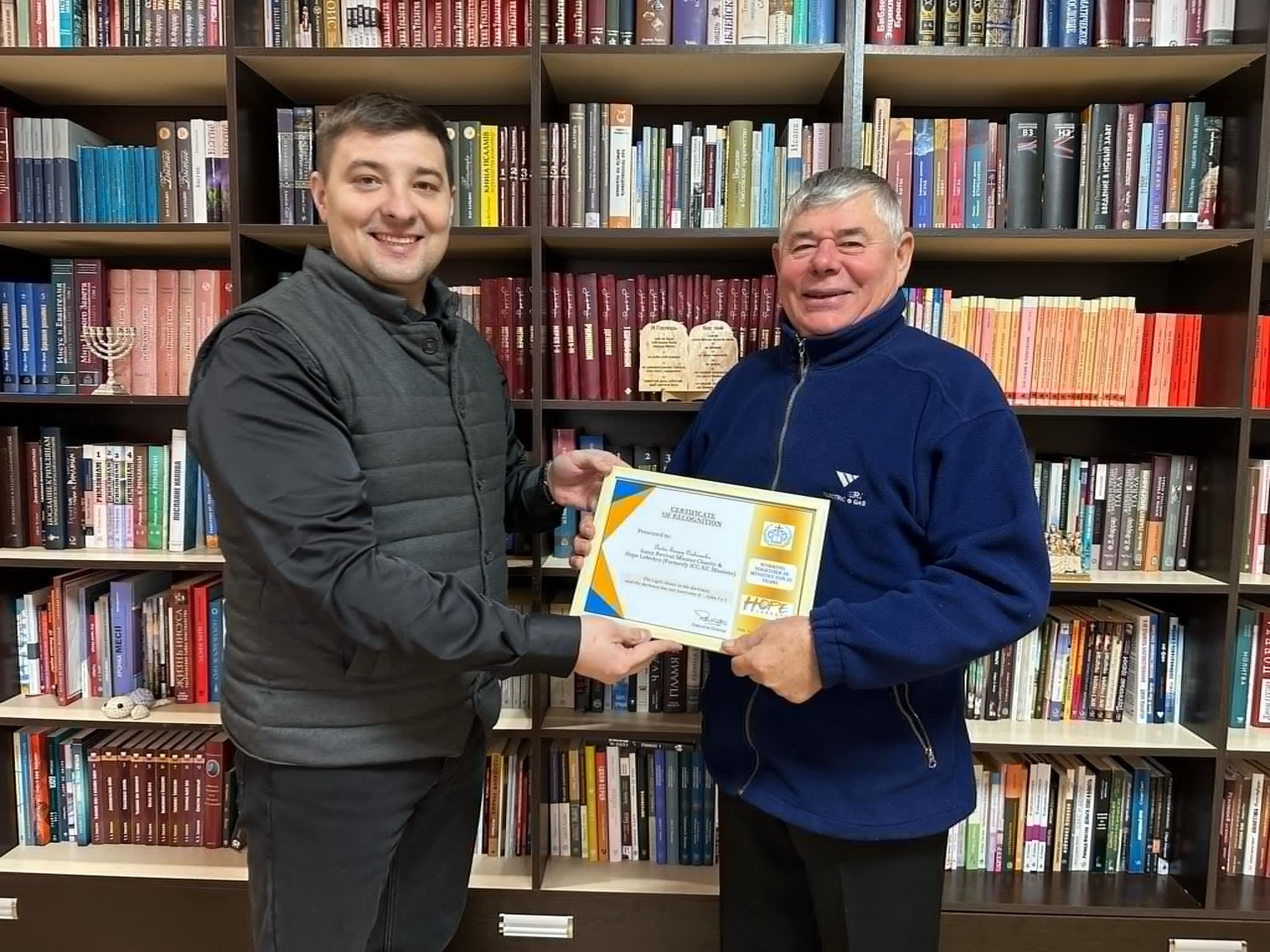 Standing in front of a bookcase, Bohdan presents Pastor Sergey with a framed certificate commemorating 25 years since Peter first met him