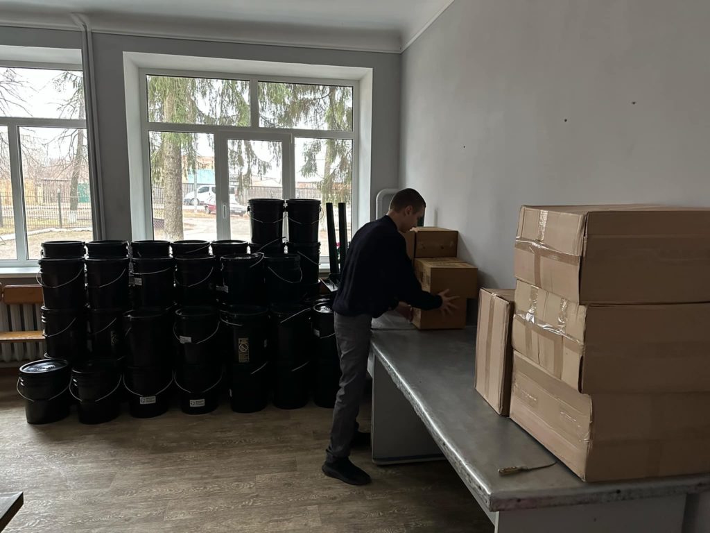 A man stacking boxes of blankets on tables, with stacks of hygiene buckets in the background