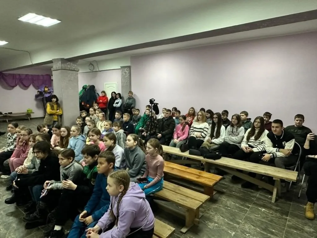 An underground bomb shelter with a low ceiling, full of children sitting on benches looking towards the puppet theatre show out of shot