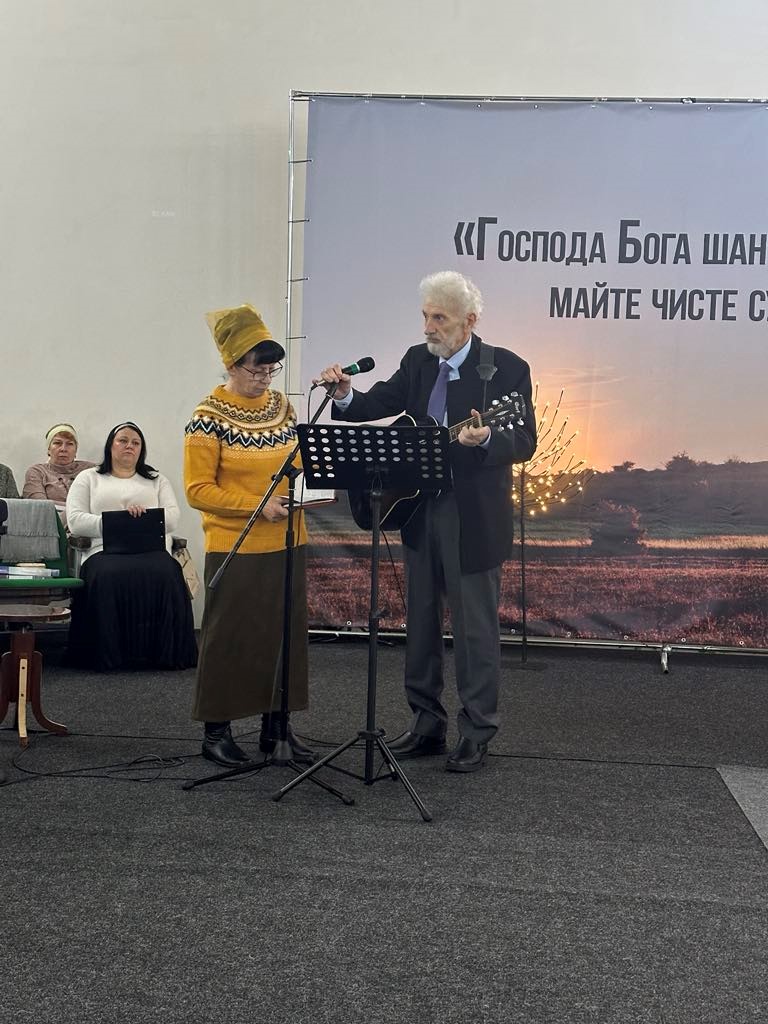 Vladimir holding a guitar and standing behind a music stand, speaking into a microphone