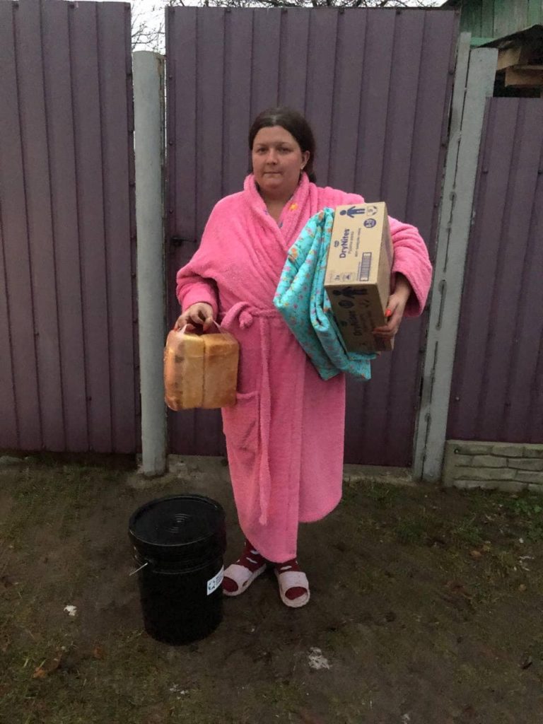 A lady standing in her dressing gown outside holding bags and boxes of donated aid