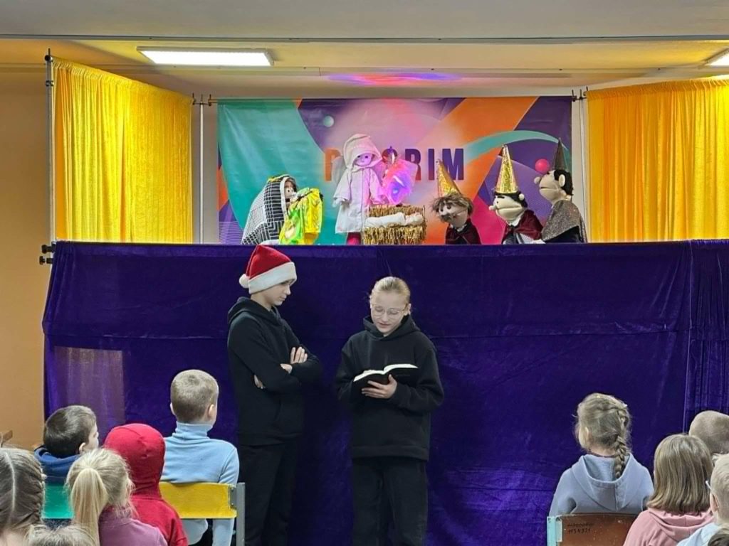 A girl reading from the Bible whilst standing in front of the puppet theatre with puppets performing behind her.