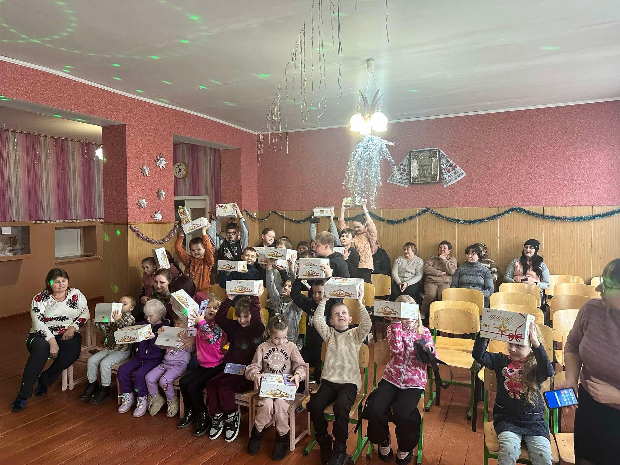 Children at the orphanage sitting and standing with their present boxes held over their heads with adults sat behind them watching