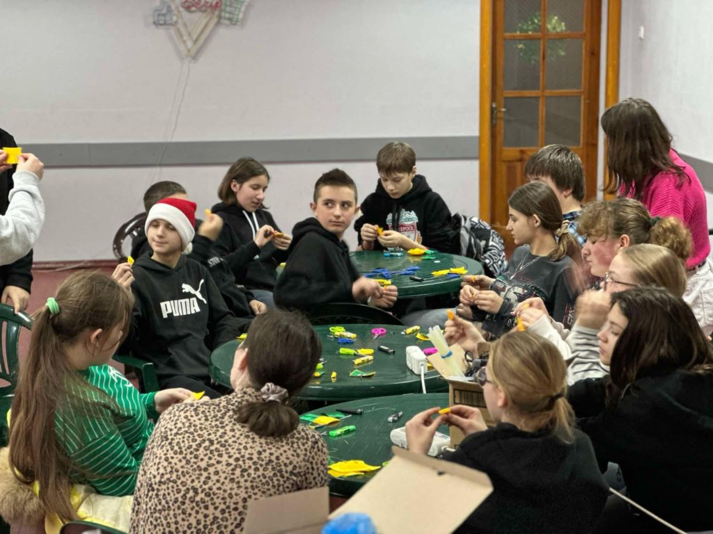 Children during the winter camp sitting at three round plastic garden tables doing crafts, with some looking over their shoulders at something out of shot