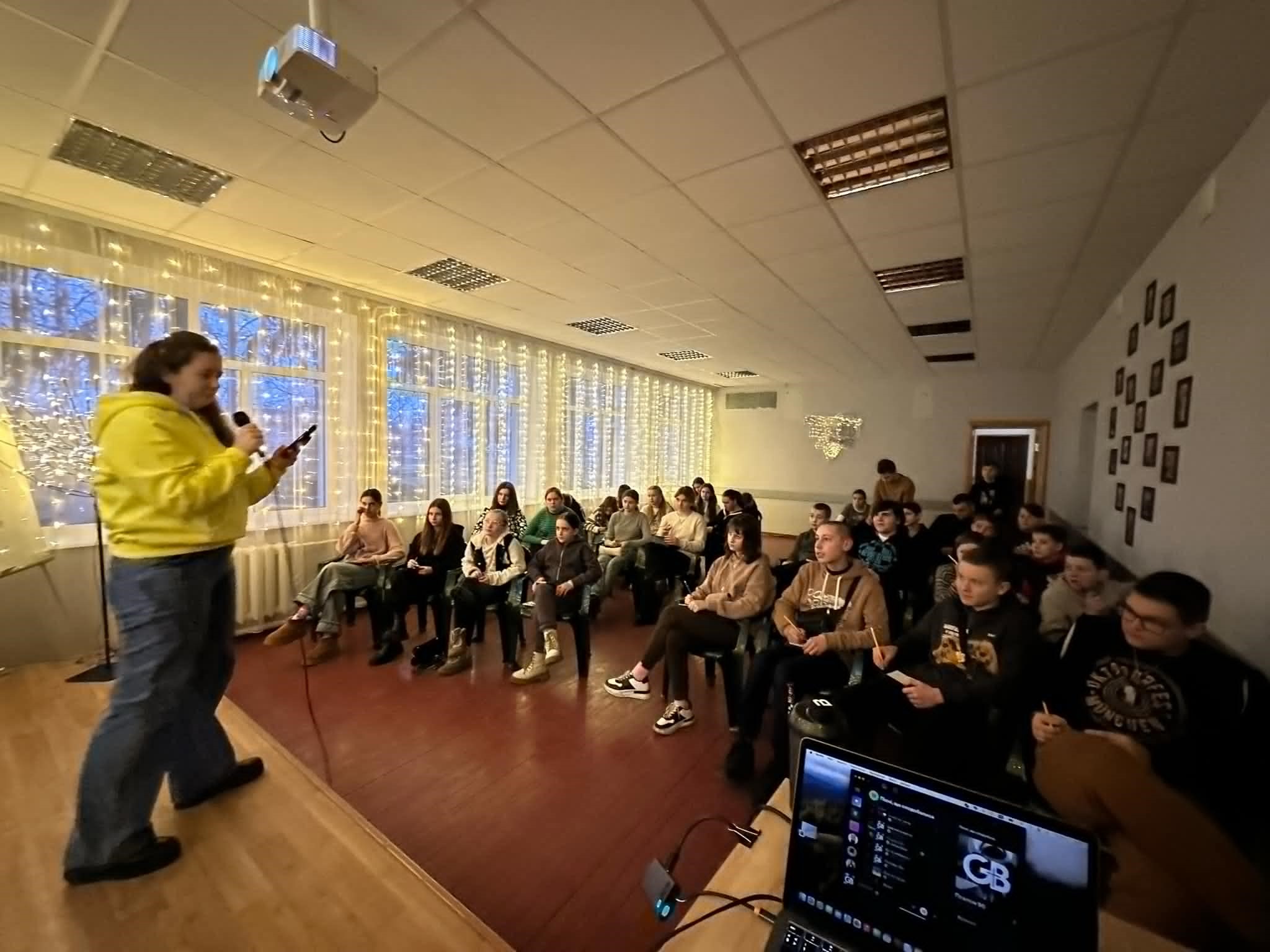 A teenage girl on a stage in the hall with other teenagers sitting listening to her speak