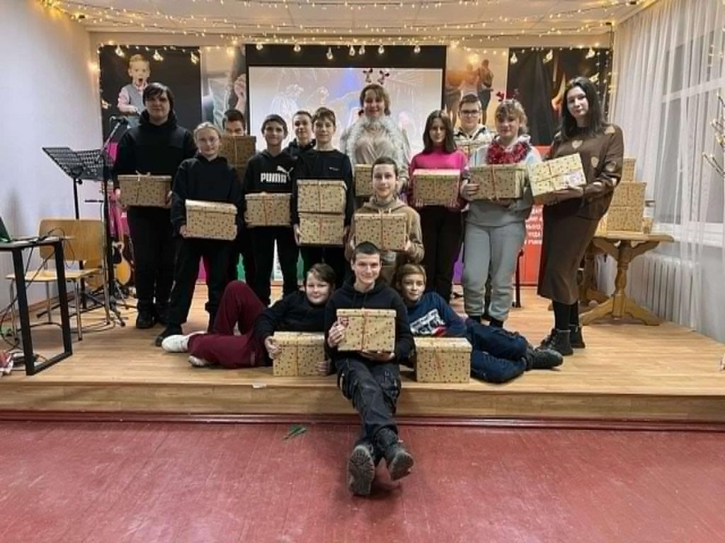 15 of the youth standing and sitting on the stage at the Centre holding Christmas presents they have been given.