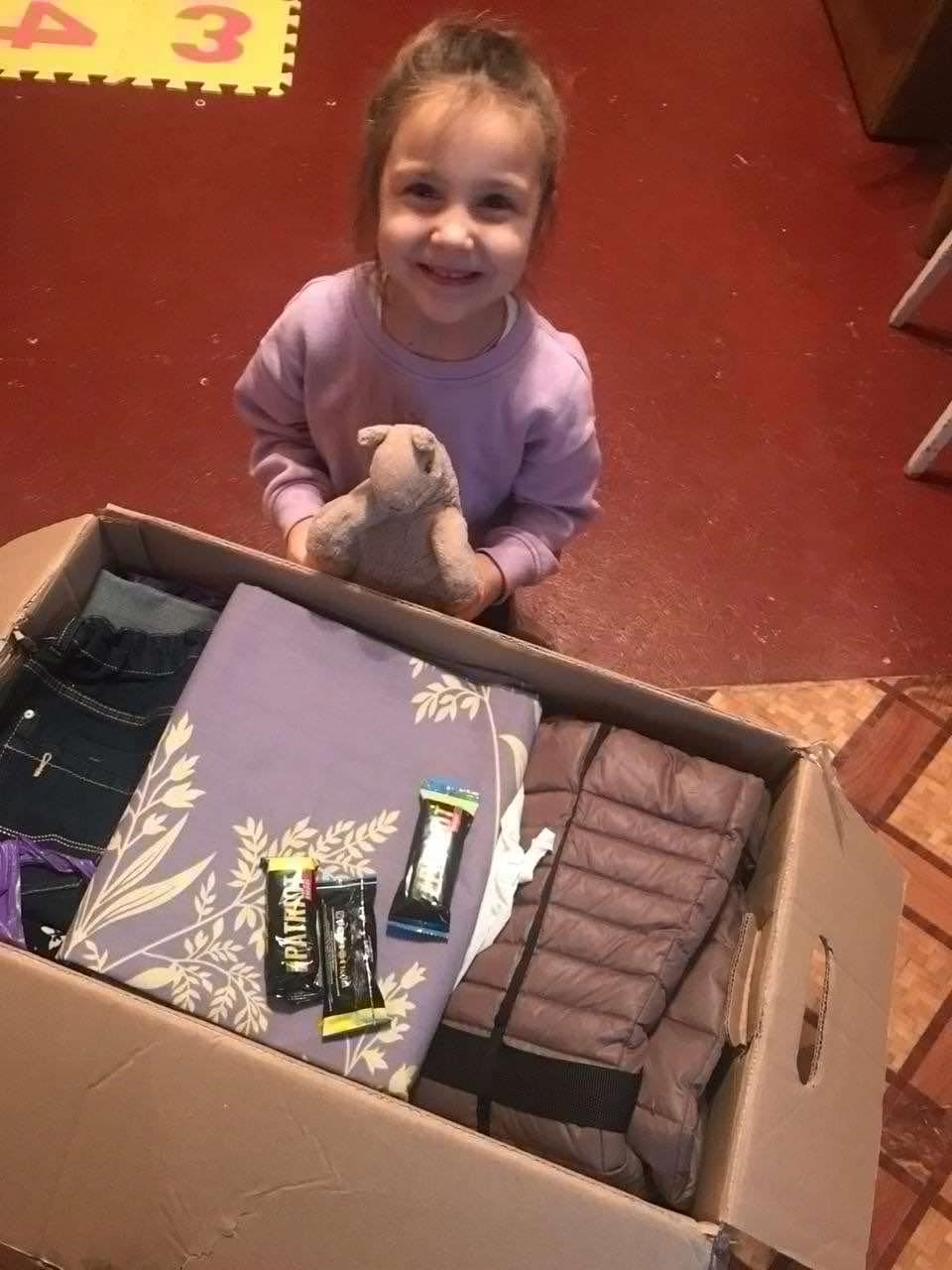 A small girl holding a cuddly toy standing behind a box of aid looking up at the camera smiling