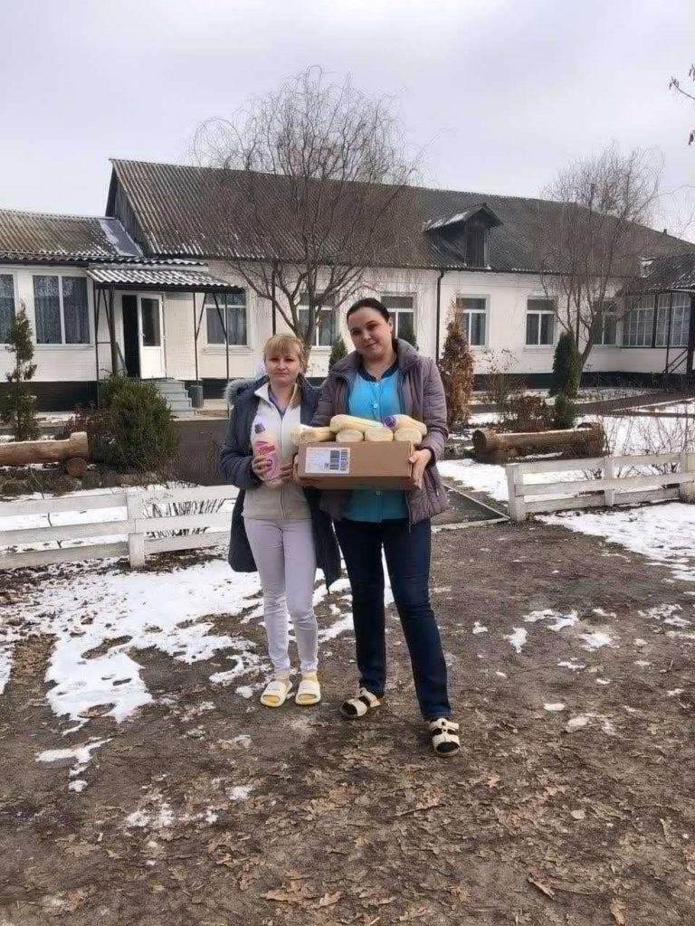 Two medical staff standing outside a medical centre in the snow holding bottles of nutrient drinks that have been donated