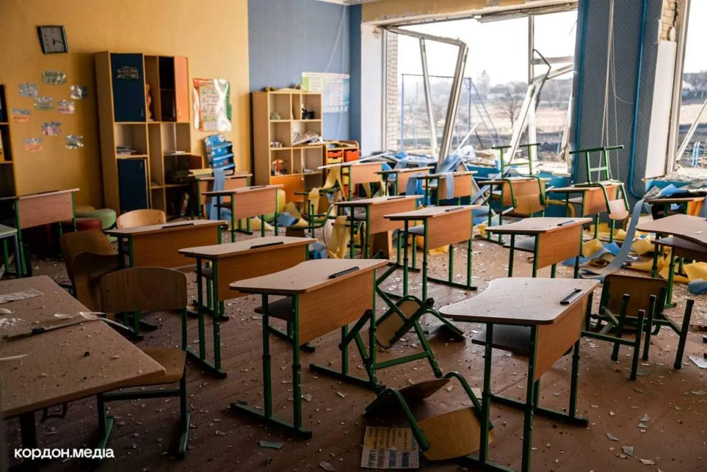 Rows of desks and chairs in a classroom cover in rubble and dust after a bomb strike with the window frames blown out.