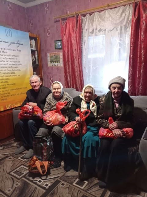 4 elderly people sat on a sofa in a living room holding bags of donated food