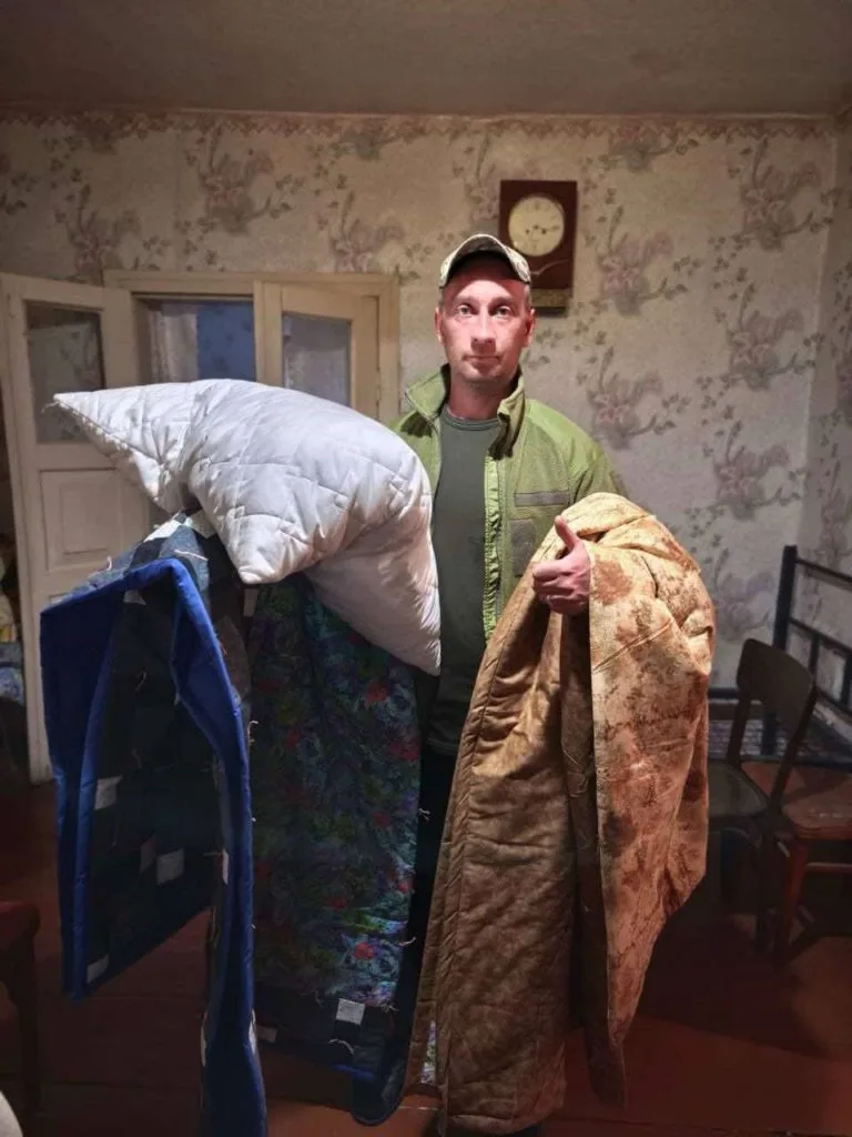 A man stood in a room of his house holding a donated pillow and two quilted blankets