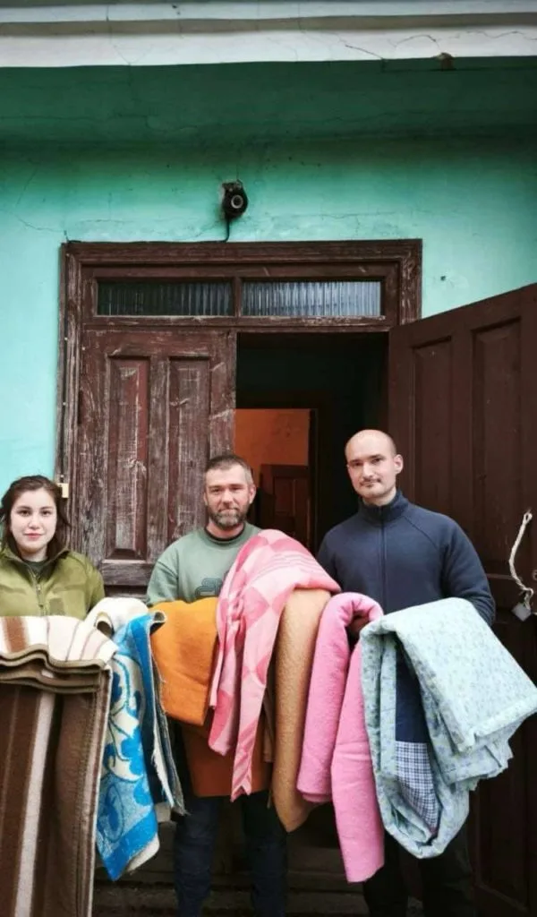 Two men and a woman standing in an old wooden doorway holding a variety donated blankets and bedding