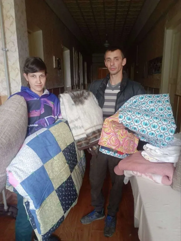 A man and a woman in their 30s standing in the hallway of a building holding a variety of donated bedding