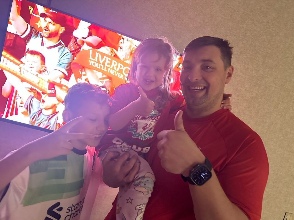 Bohdan standing in front of a TV showing celebrating Liverpool fans after they won the Premier League title