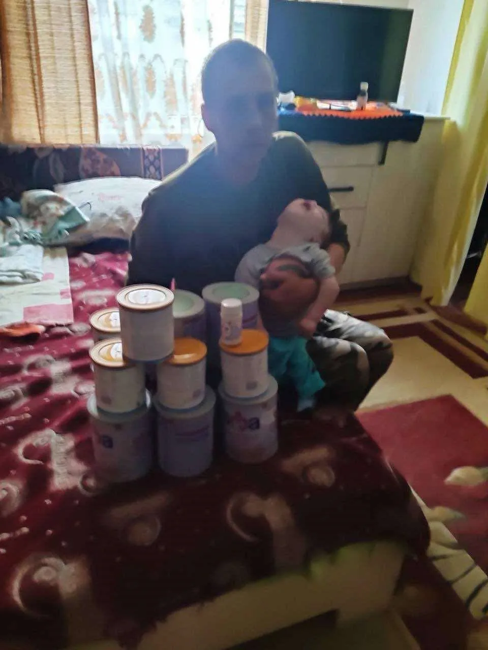 A father sitting on a bed cradling his young child with tins of special baby milk stacked on the bed in front of him