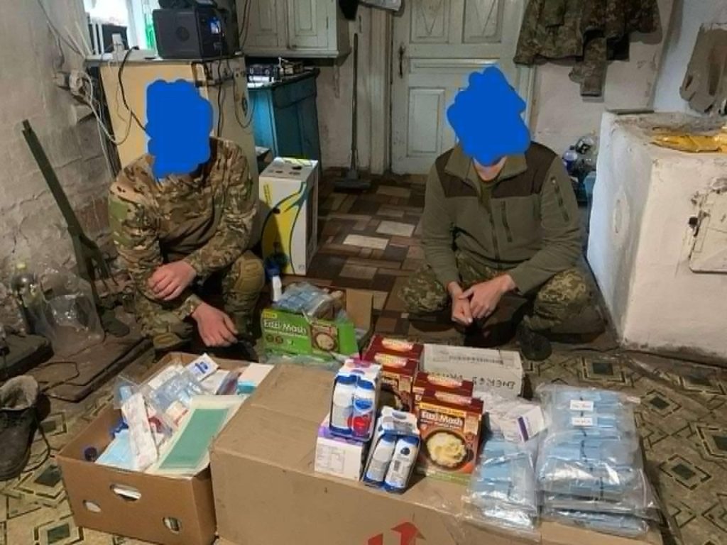 Two soldiers with thei faces obscured, sat cross legged on the floor behind boxes of aid sent by the church