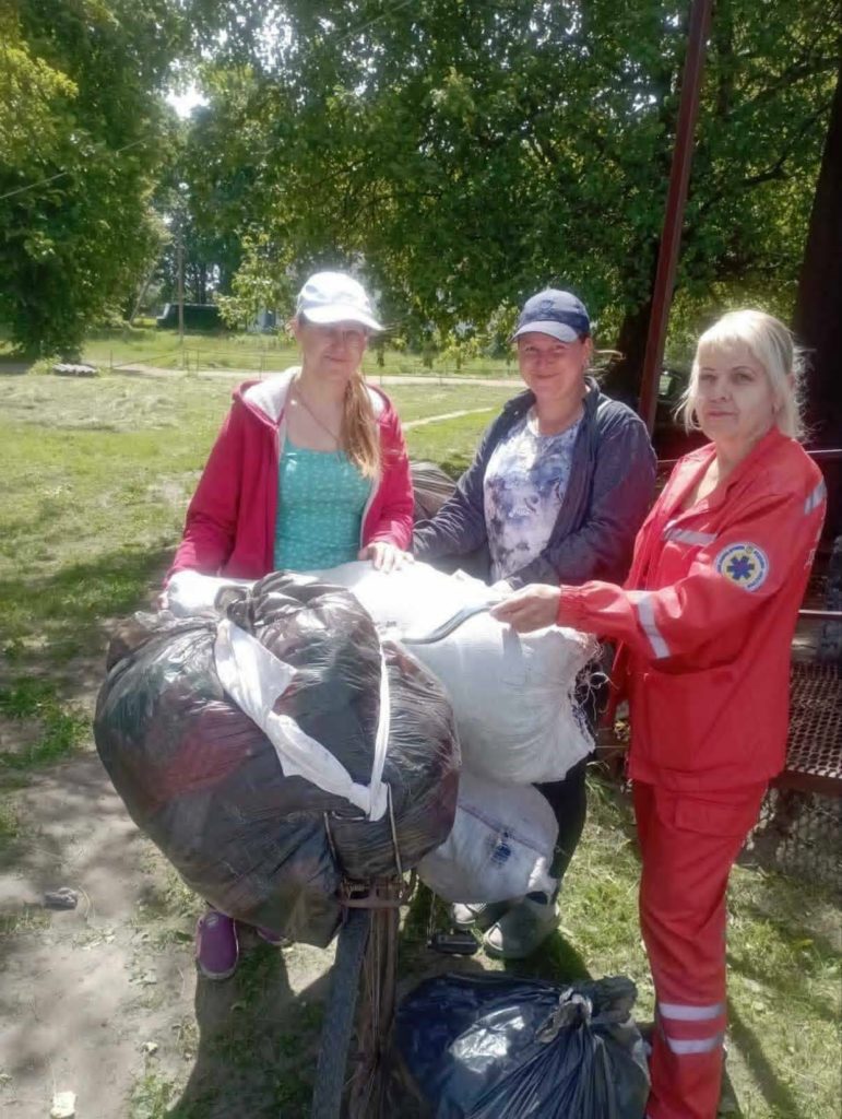 Three ladies standing around a bicycle that has bags of clothes piled on the front.