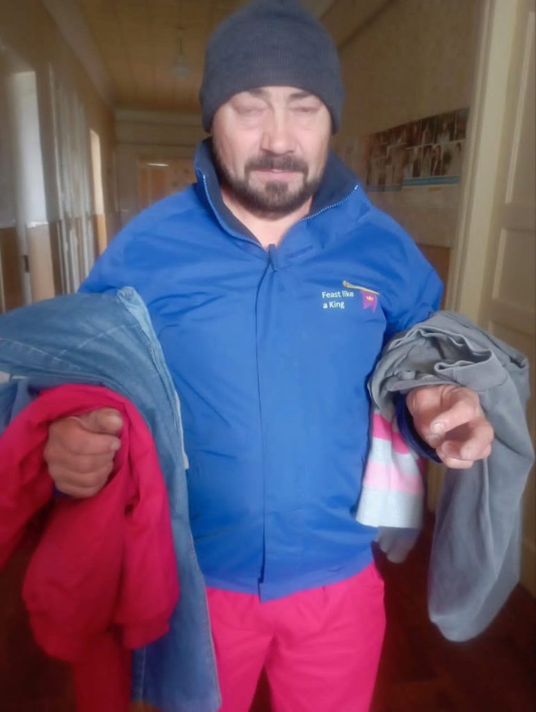 A man wearing a beanie hat standing in a corridor with donated clothes draped over his arms