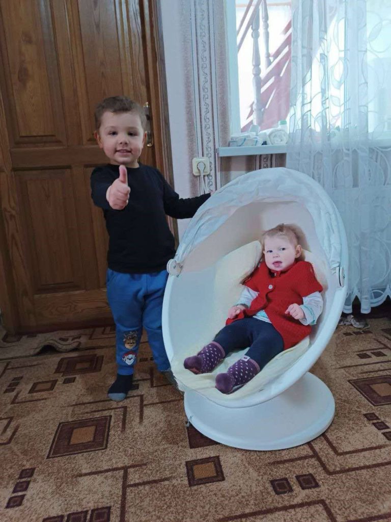 A toddler sat in a small egg chair with a young boy standing next to her giving a thumbs up