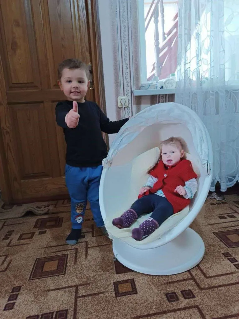 A toddler sat in a small egg chair with a young boy standing next to her giving a thumbs up