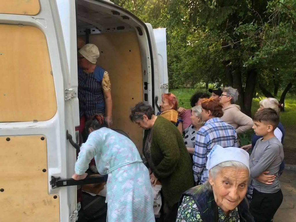 Group of people, including elderly women and children, gathered around the open back of a van where a woman is distributing loaves of bread in an outdoor setting with trees in the background.