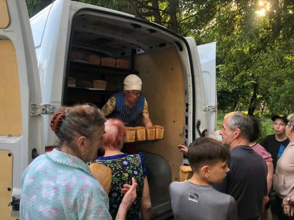 Woman handing out loaves of bread from the back of a van to a gathered crowd of people, including elderly individuals and children, in a wooded outdoor setting during daylight.
