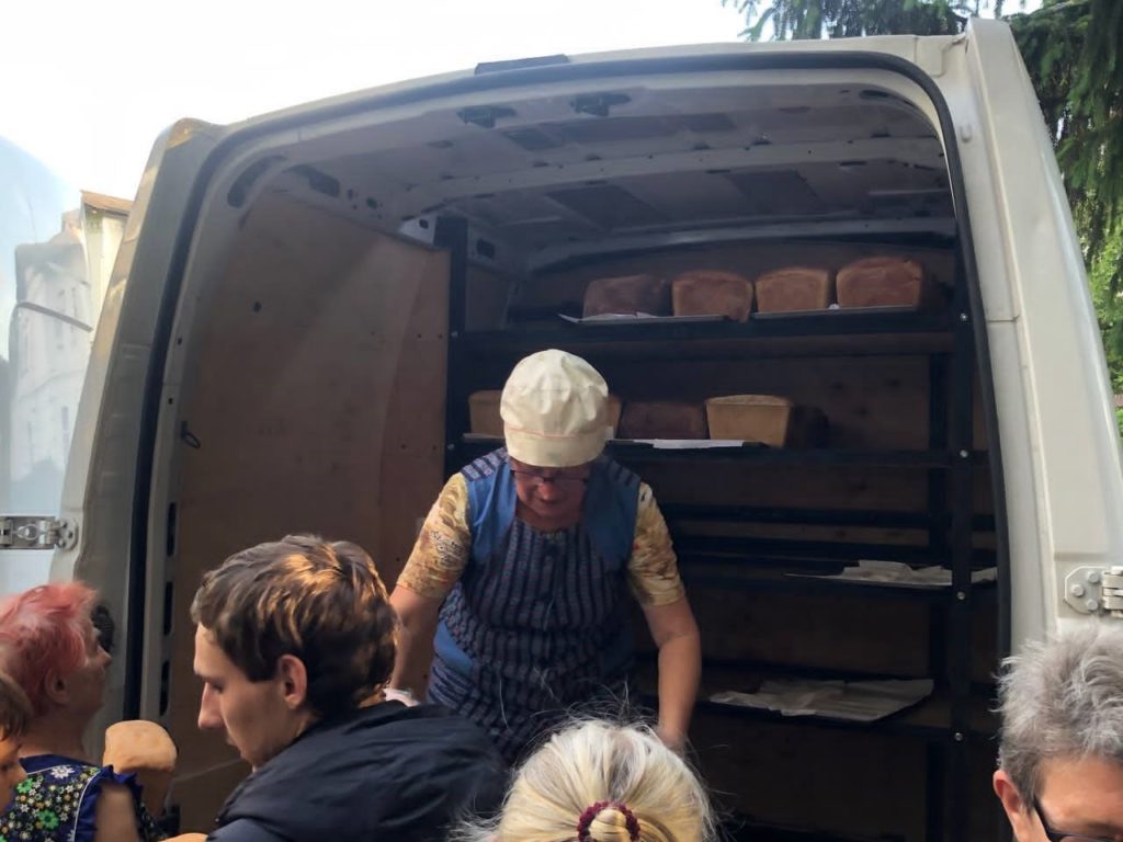 Person distributing fresh loaves of bread from the back of a van to a group of people, with shelves inside the van stocked with bread and individuals reaching out to receive it.