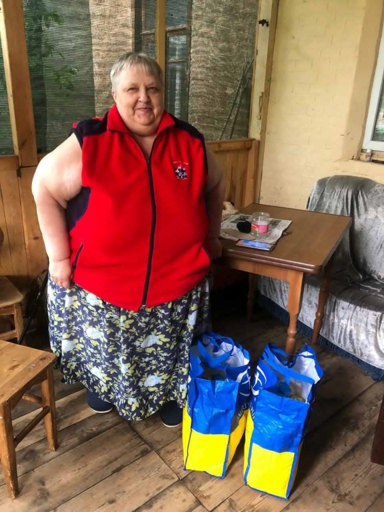 Smiling woman standing indoors next to two large blue and yellow shopping bags filled with supplies, in a rustic room with wooden floors, simple furniture, and a table with newspapers and a candle.